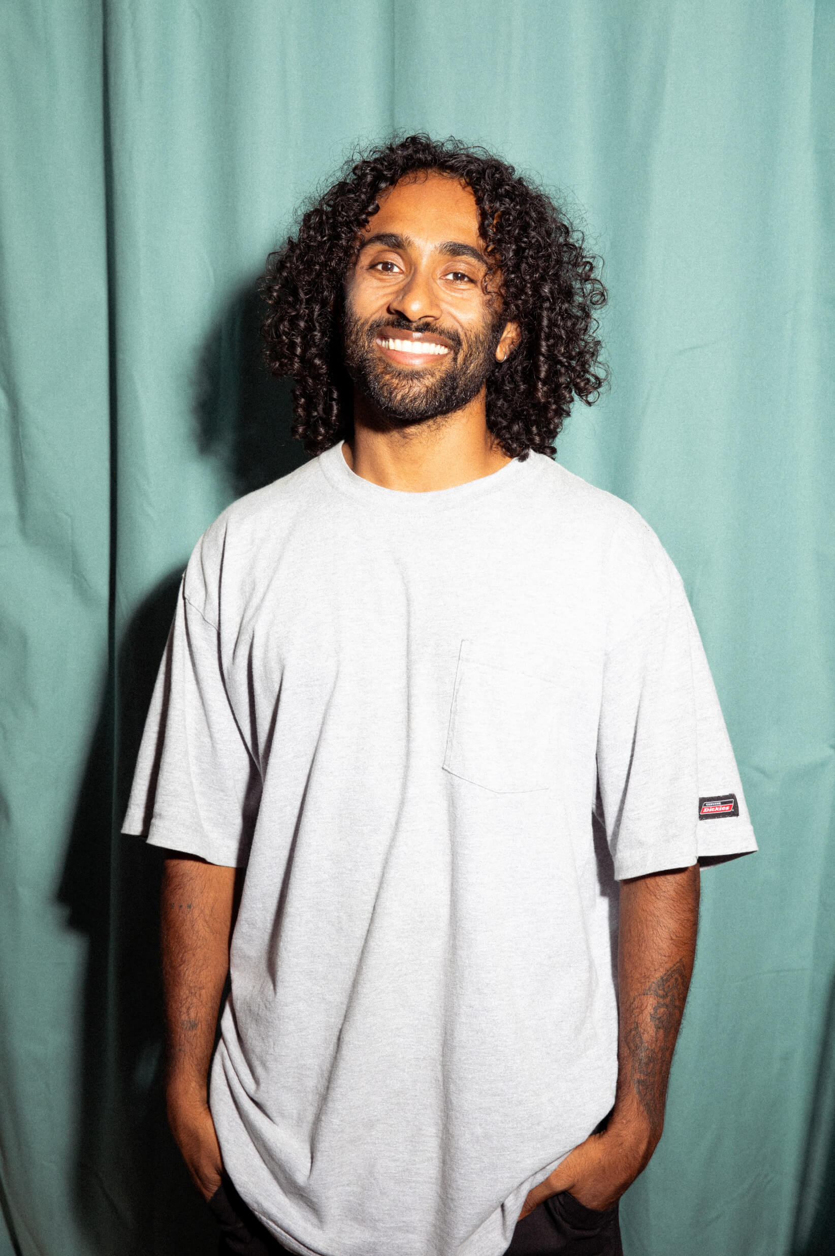 A man with long curly dark hair and a beard smiles at the camera. He's wearing a white t-shirt and standing in front of a light teal backdrop.