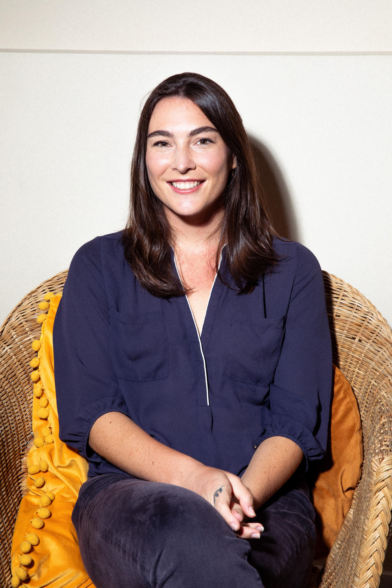 A smiling woman with long dark hair sits in a wicker chair. She's wearing a navy blue top and there's a yellow cushion or blanket visible behind her.